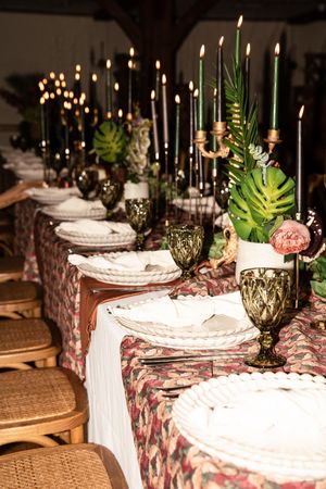 Elegant wedding reception table with dark candles, tropical greenery, floral tablecloth, and green goblets.