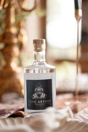 A bottle of King Arthur Premium Gin with cork stopper sits on a table beside a champagne flute in warm golden light.