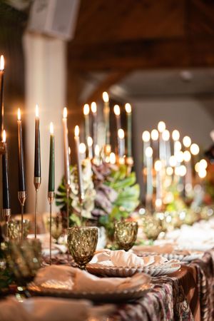 Romantic candlelit reception table with tall taper candles, green glassware, tropical florals, and warm ambient lighting.