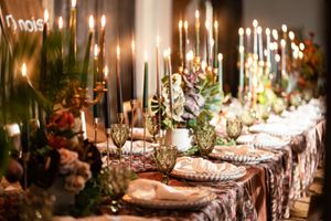 Elegant wedding reception table with tall candles, lush floral centerpieces, green goblets, and patterned linens in warm lighting.