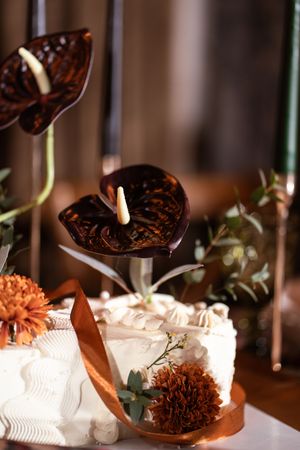 Elegant white wedding cake decorated with dark chocolate anthurium flowers, orange chrysanthemums, and a satin ribbon.