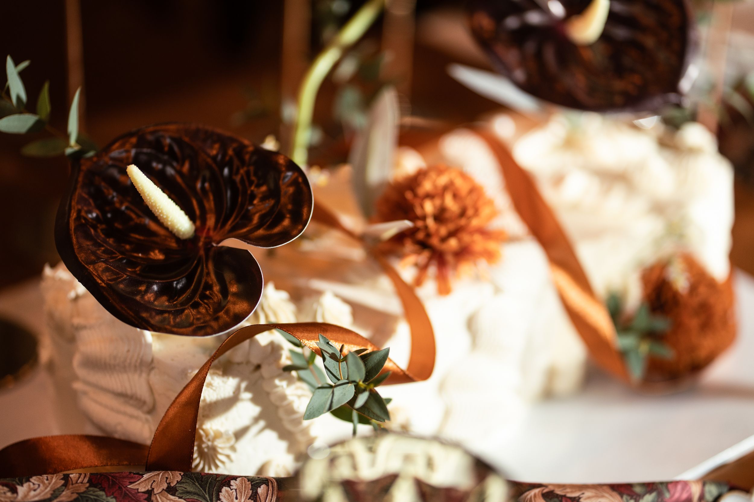 Elegant wedding cake display with chocolate decorations, burnt orange ribbons, and floral accents in warm autumn tones.