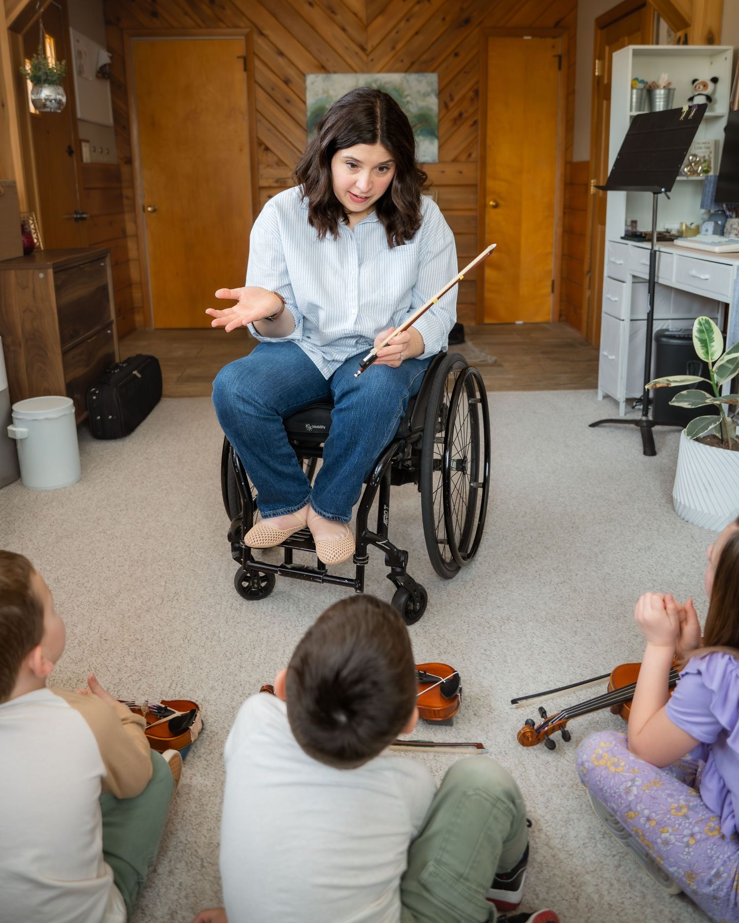 A woman in a wheelchair holds a baton while teaching violin to children sitting on the floor in a cozy home.