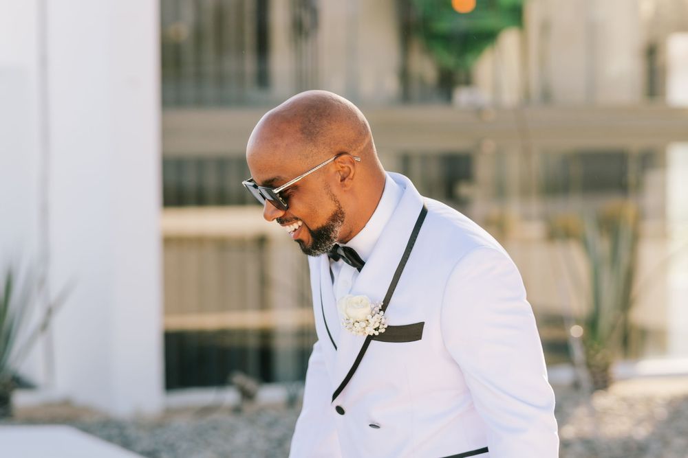 Bald groom in white tuxedo jacket with bow tie and boutonniere wearing sunglasses outdoors at wedding.