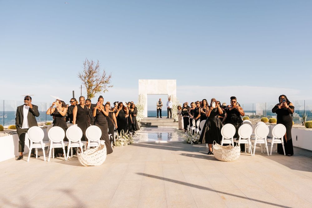 Outdoor rooftop wedding ceremony with guests in black attire, white chairs, and a minimalist arch under a clear blue sky.