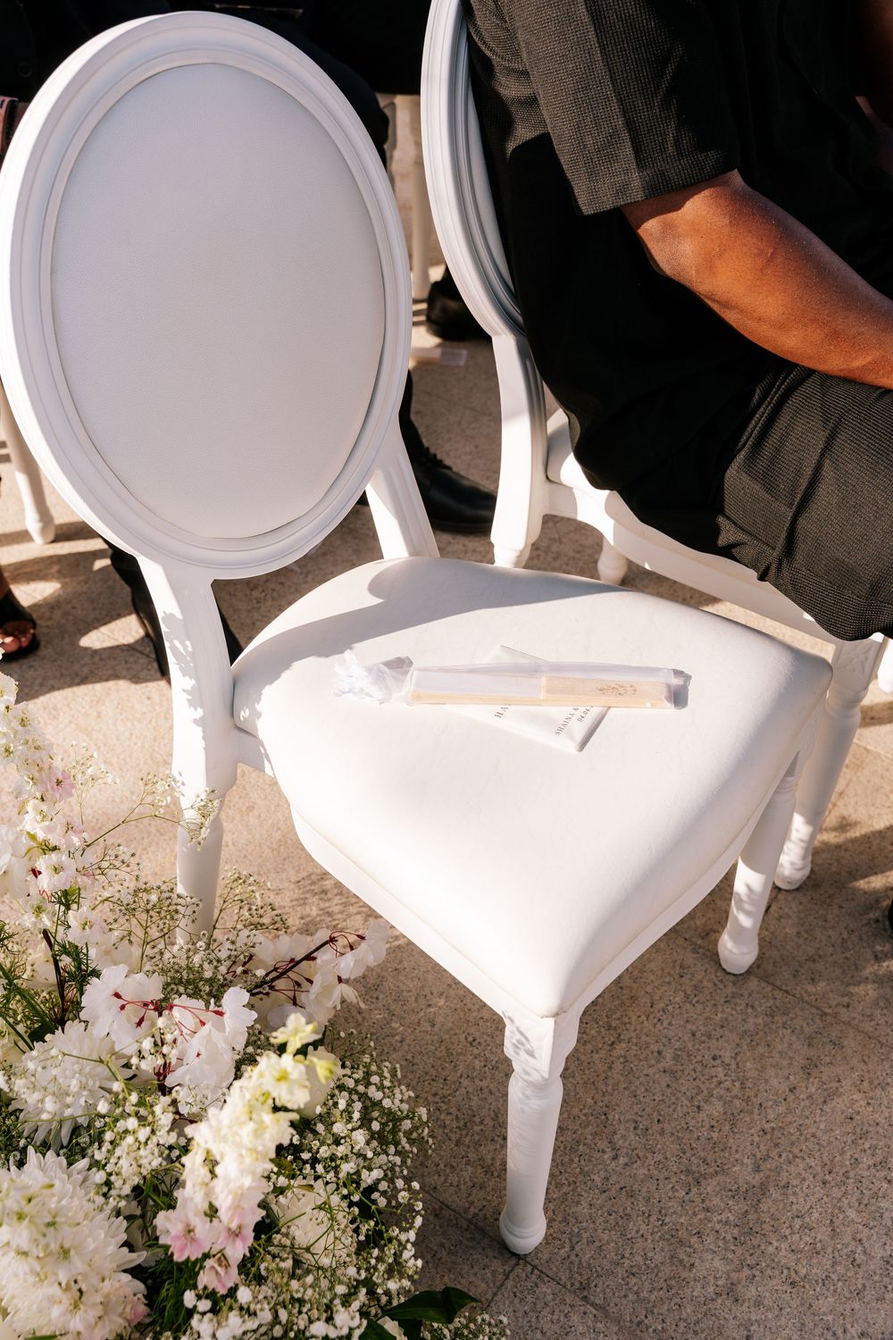 Elegant white Louis XVI chair with wedding programs on seat, surrounded by floral arrangements at an outdoor ceremony.