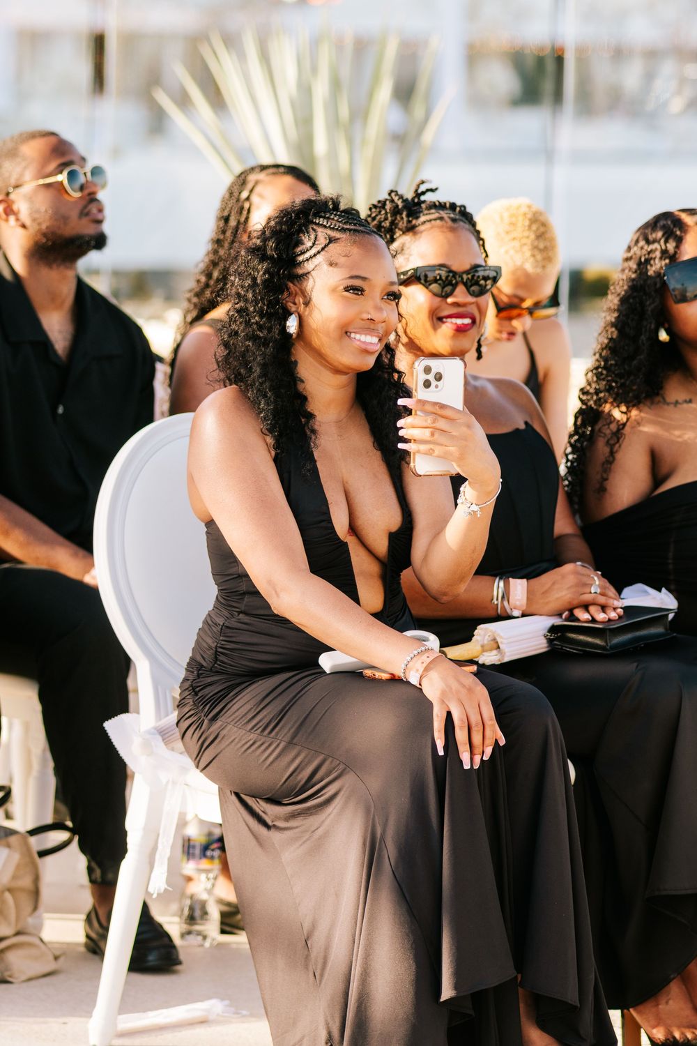 Smiling woman in black dress at outdoor wedding ceremony with guests seated behind her.