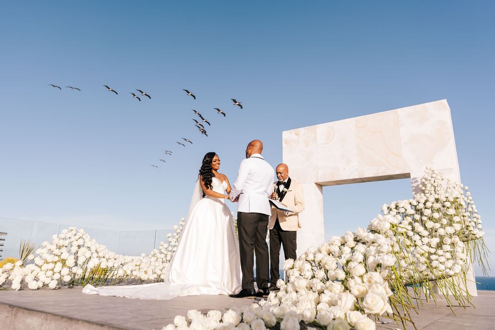Outdoor wedding ceremony with white floral arch, bride and groom exchanging vows, birds flying overhead against a clear blue sky.