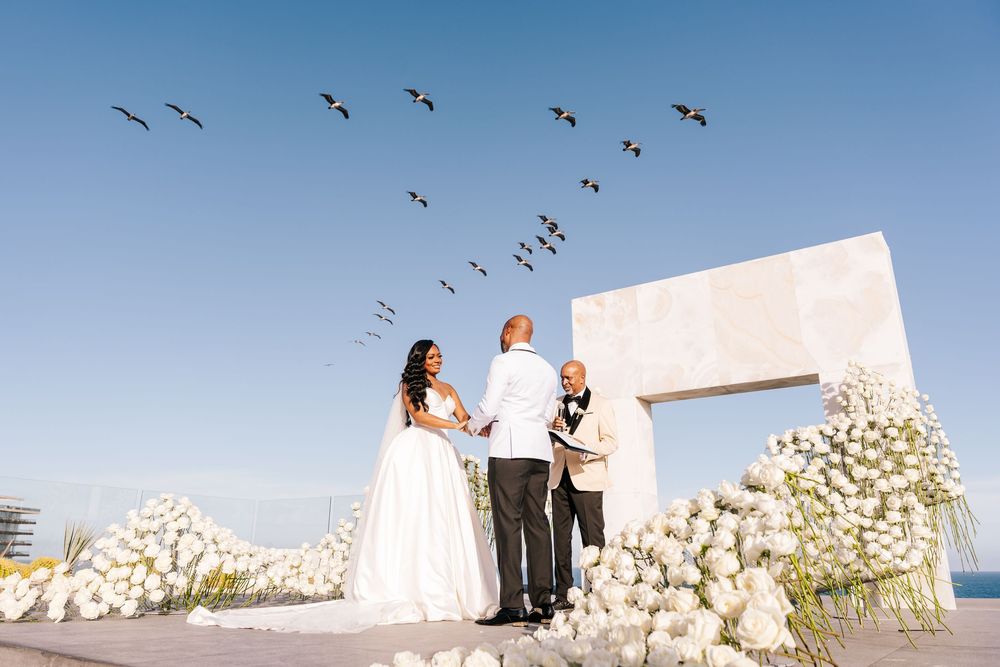 Couple exchanges vows at an elegant outdoor rooftop wedding ceremony with white floral arrangements and birds flying overhead.