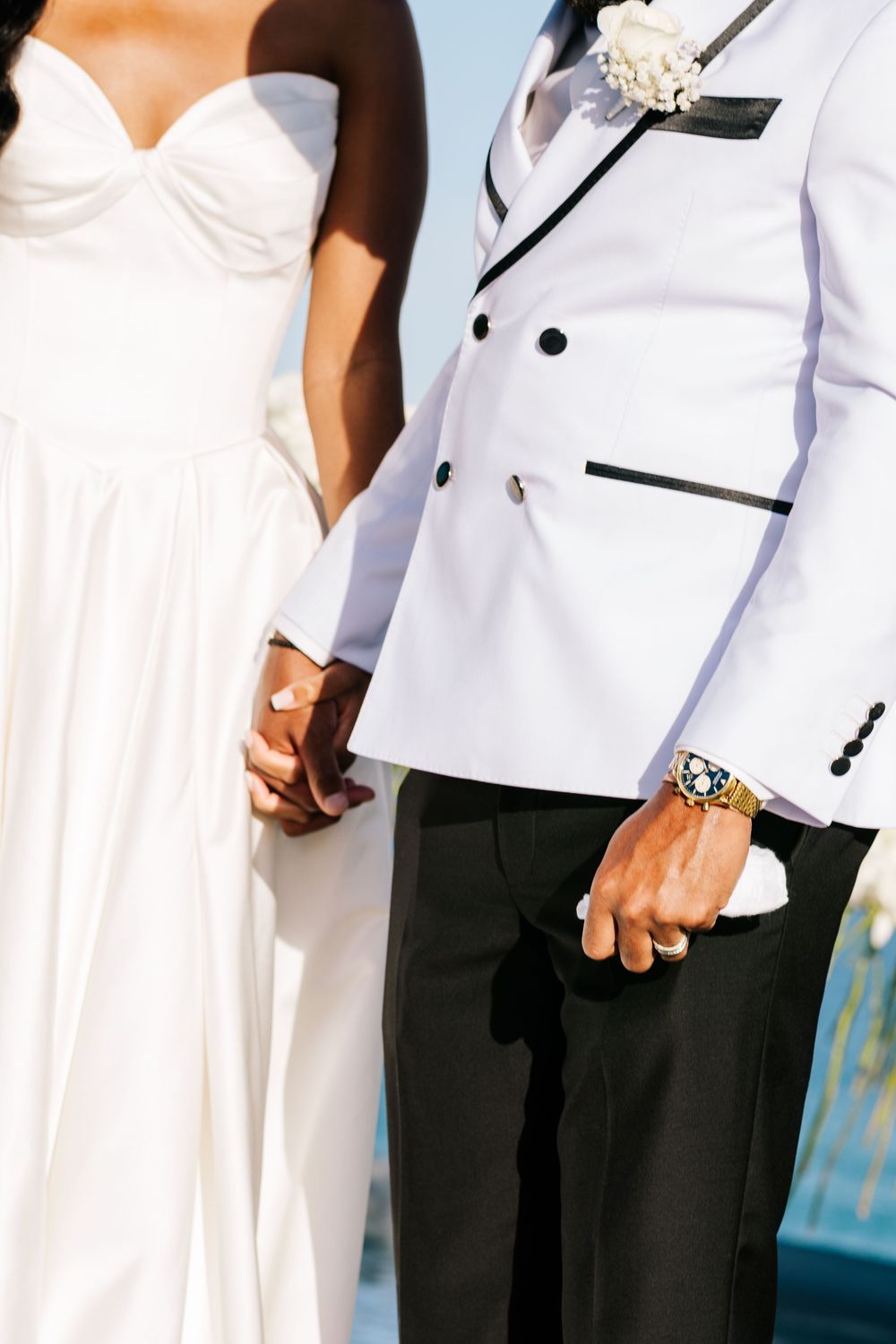 Bride and groom in white wedding attire holding hands, groom wearing a bold gold watch and white boutonniere.