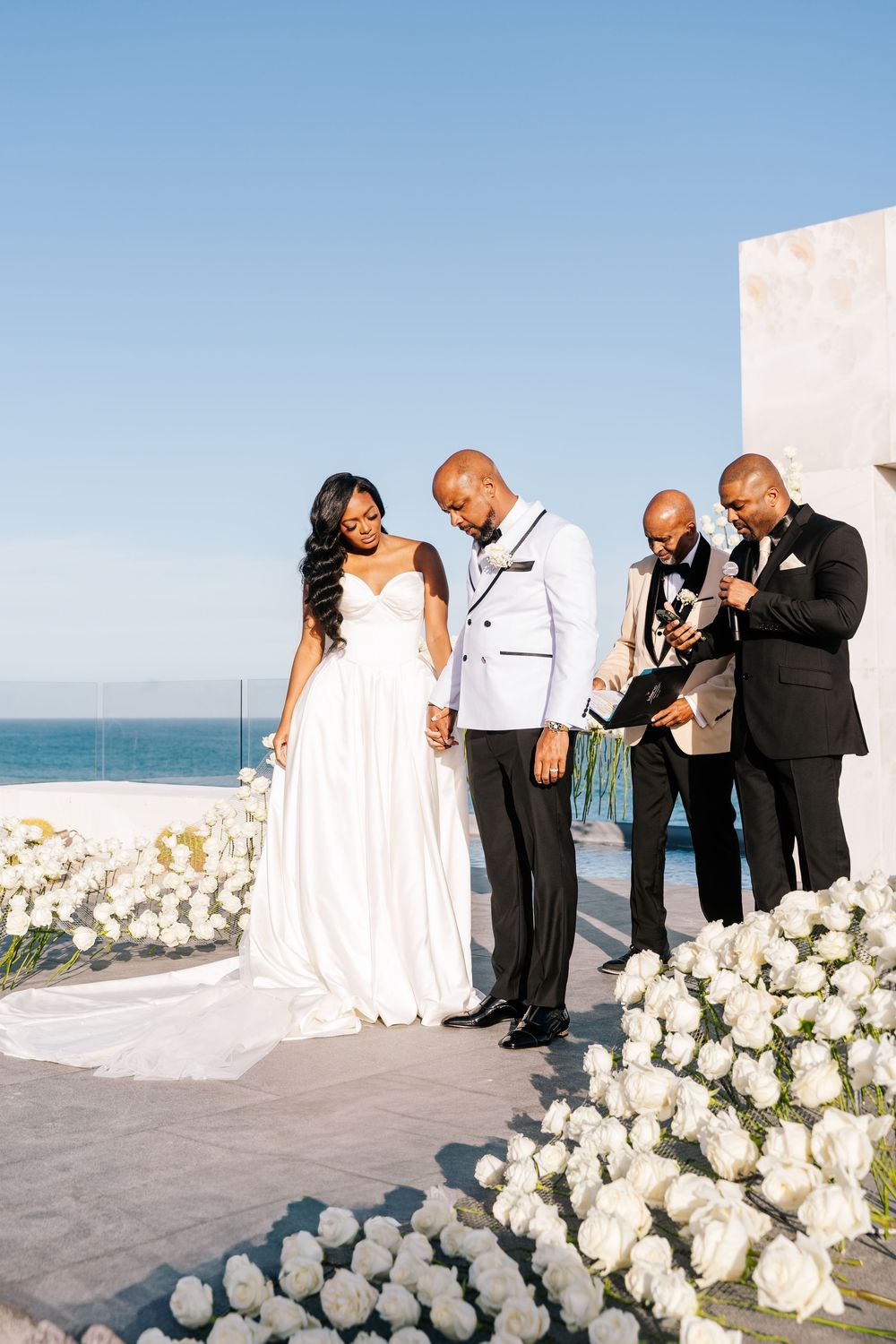 Bride and groom hold hands during outdoor wedding ceremony with white roses and ocean backdrop.