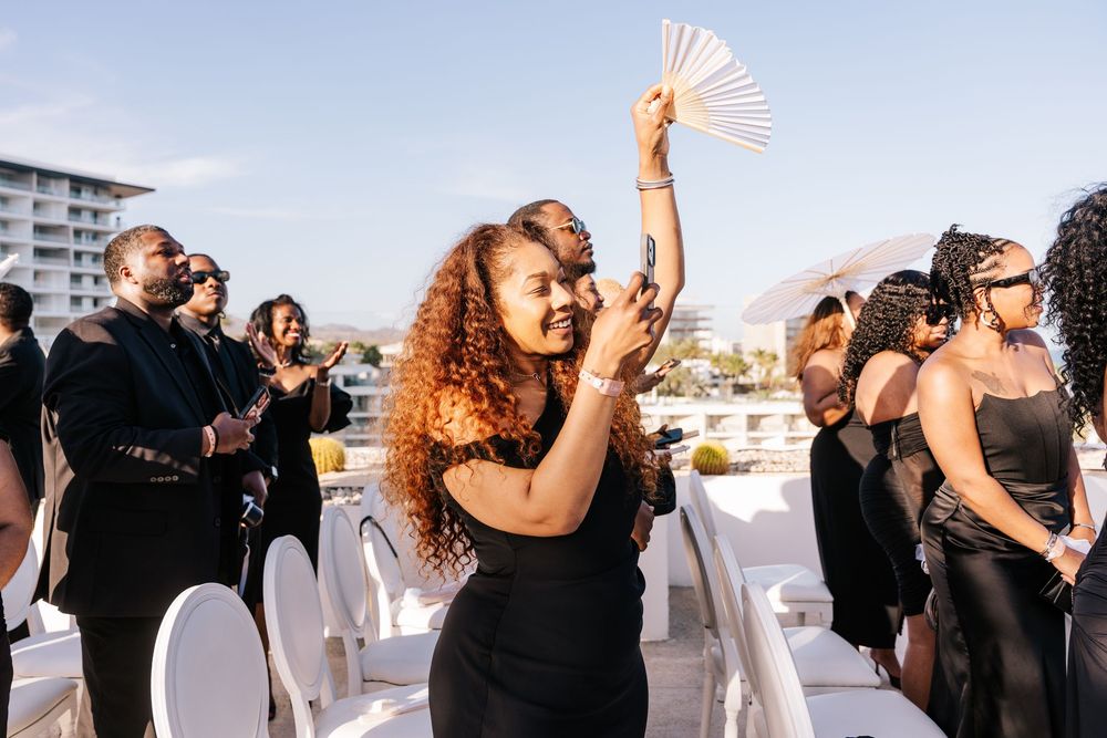 Wedding guest in black dress dancing joyfully with a hand fan at an outdoor rooftop wedding celebration.