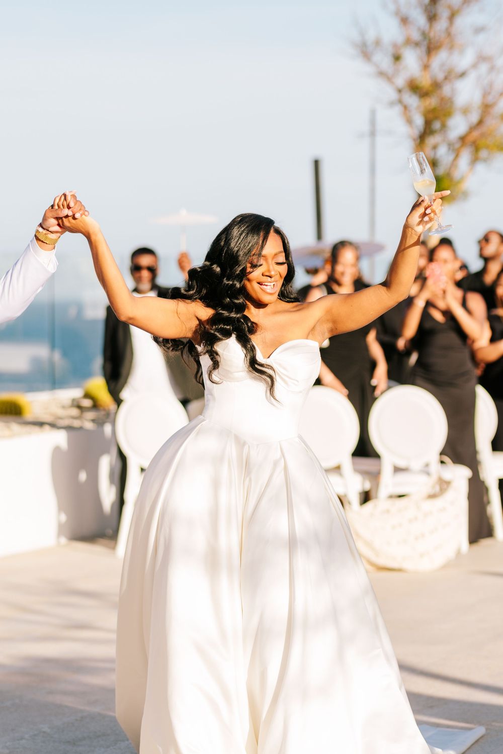 Radiant bride in a strapless white gown celebrating with arms raised and champagne glass at outdoor wedding reception.