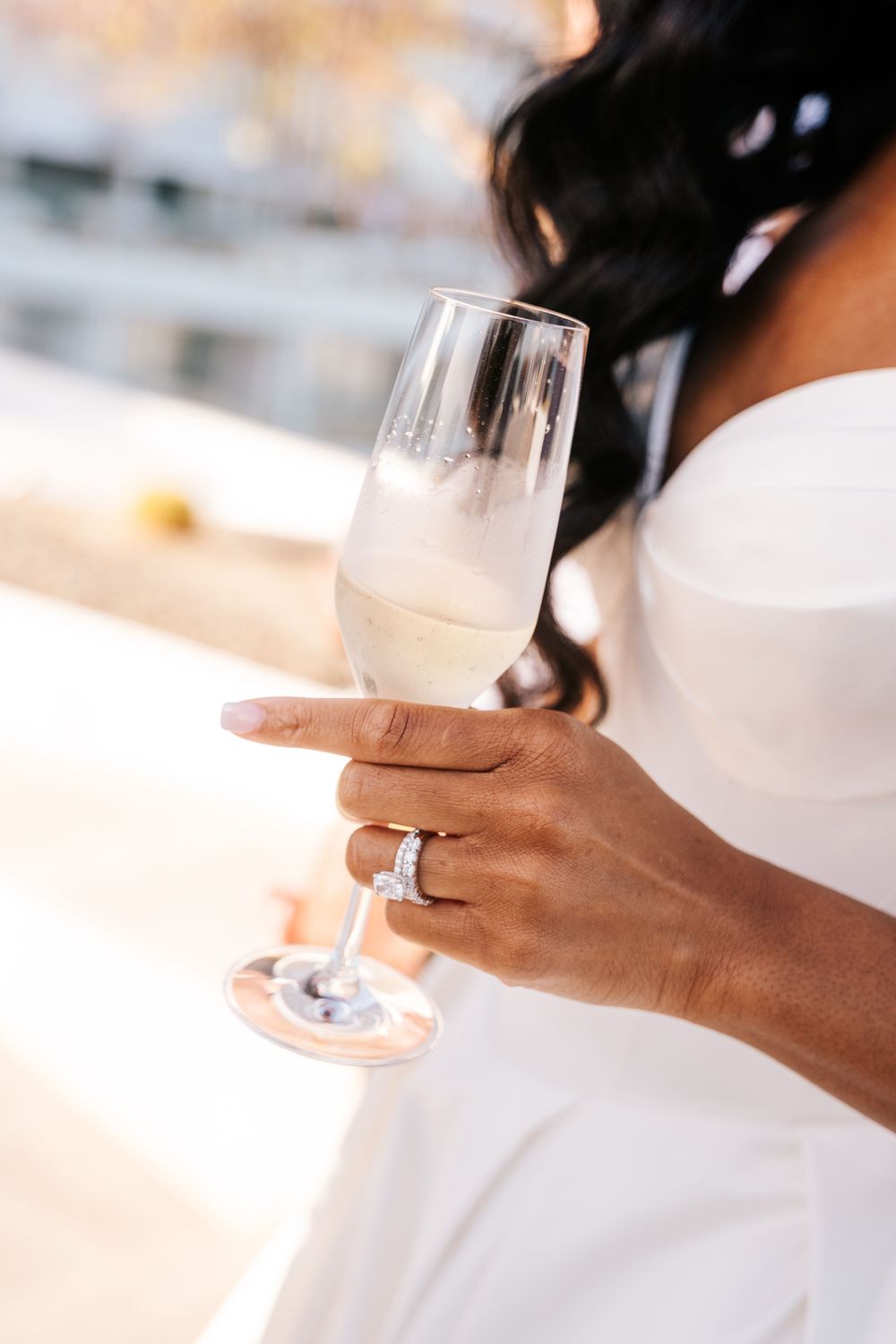 Close-up of a bride in a white strapless dress holding a champagne flute, wearing a diamond engagement ring.