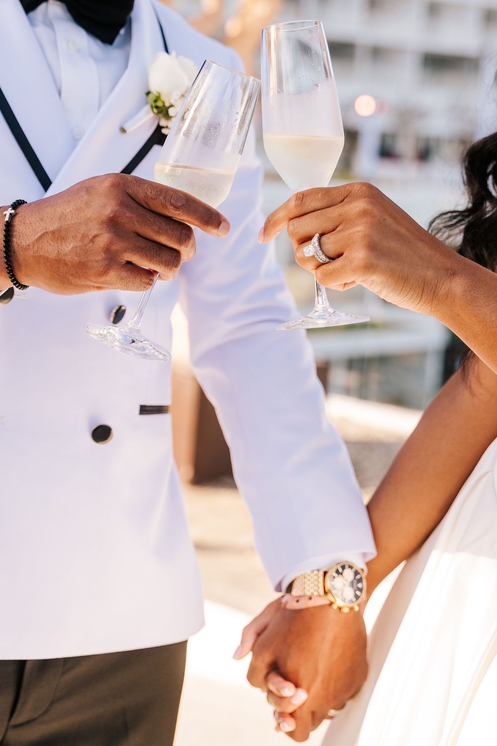 Bride and groom clinking champagne glasses while holding hands, showcasing wedding rings and a luxury gold watch.