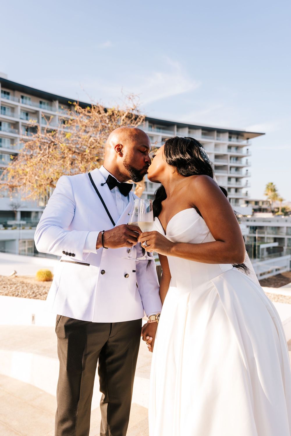 Bride and groom kissing while holding champagne glasses at outdoor wedding venue.