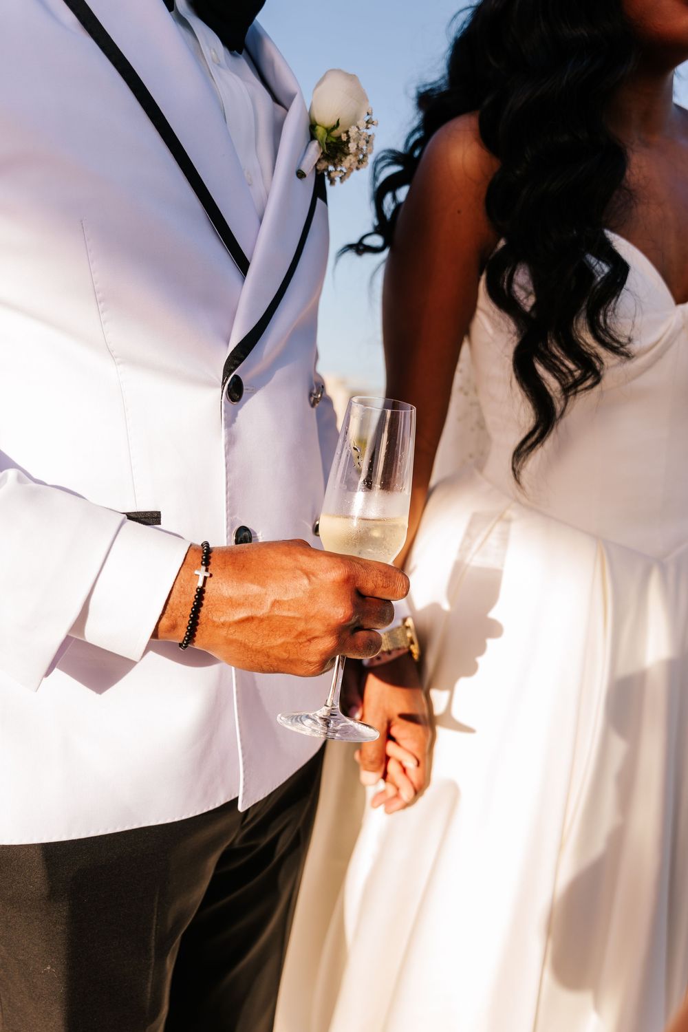 Interracial couple holding champagne flutes at outdoor wedding ceremony, groom in white suit with boutonniere.