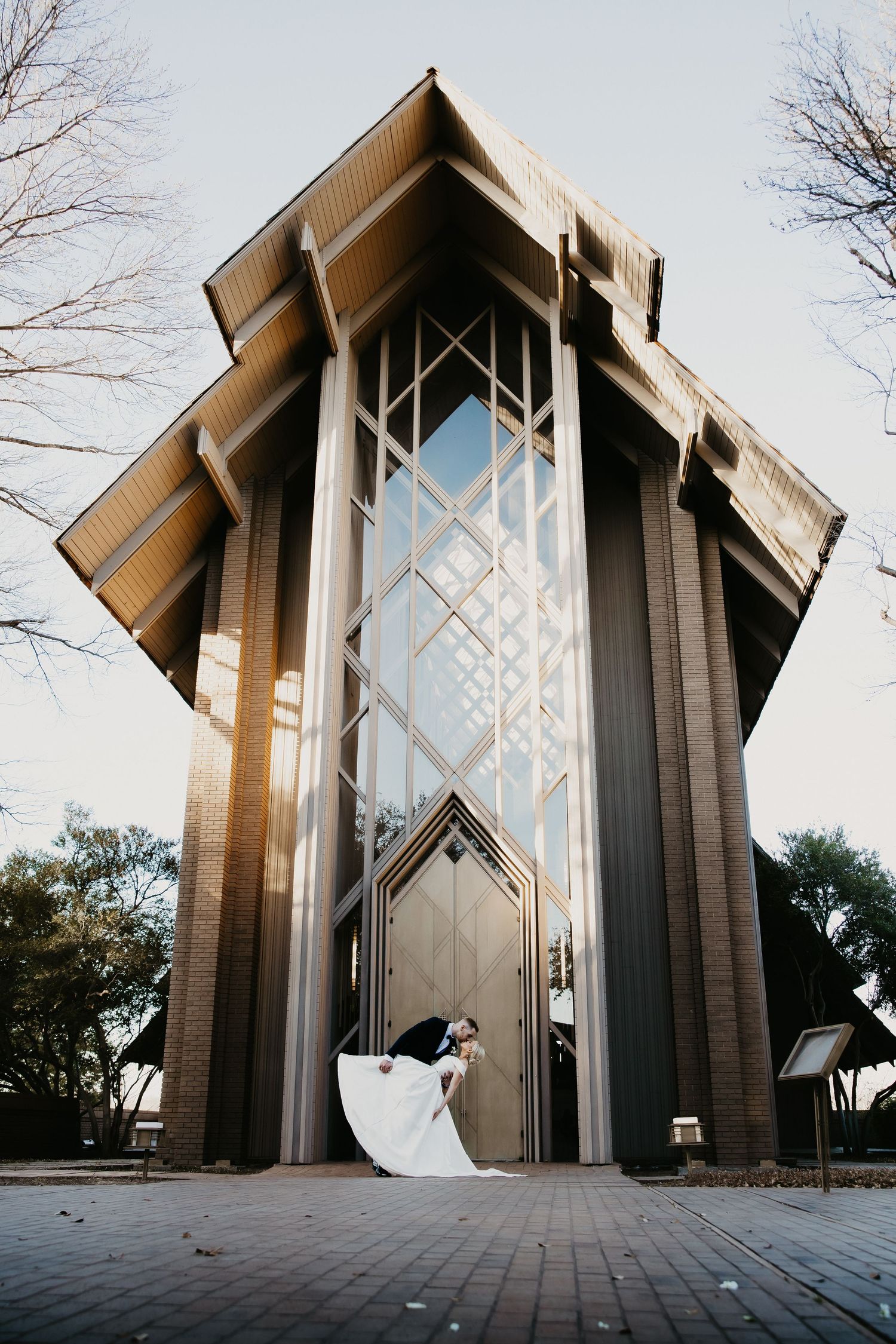 Bride and groom share a romantic dip kiss in front of a stunning modern chapel with tall glass windows.