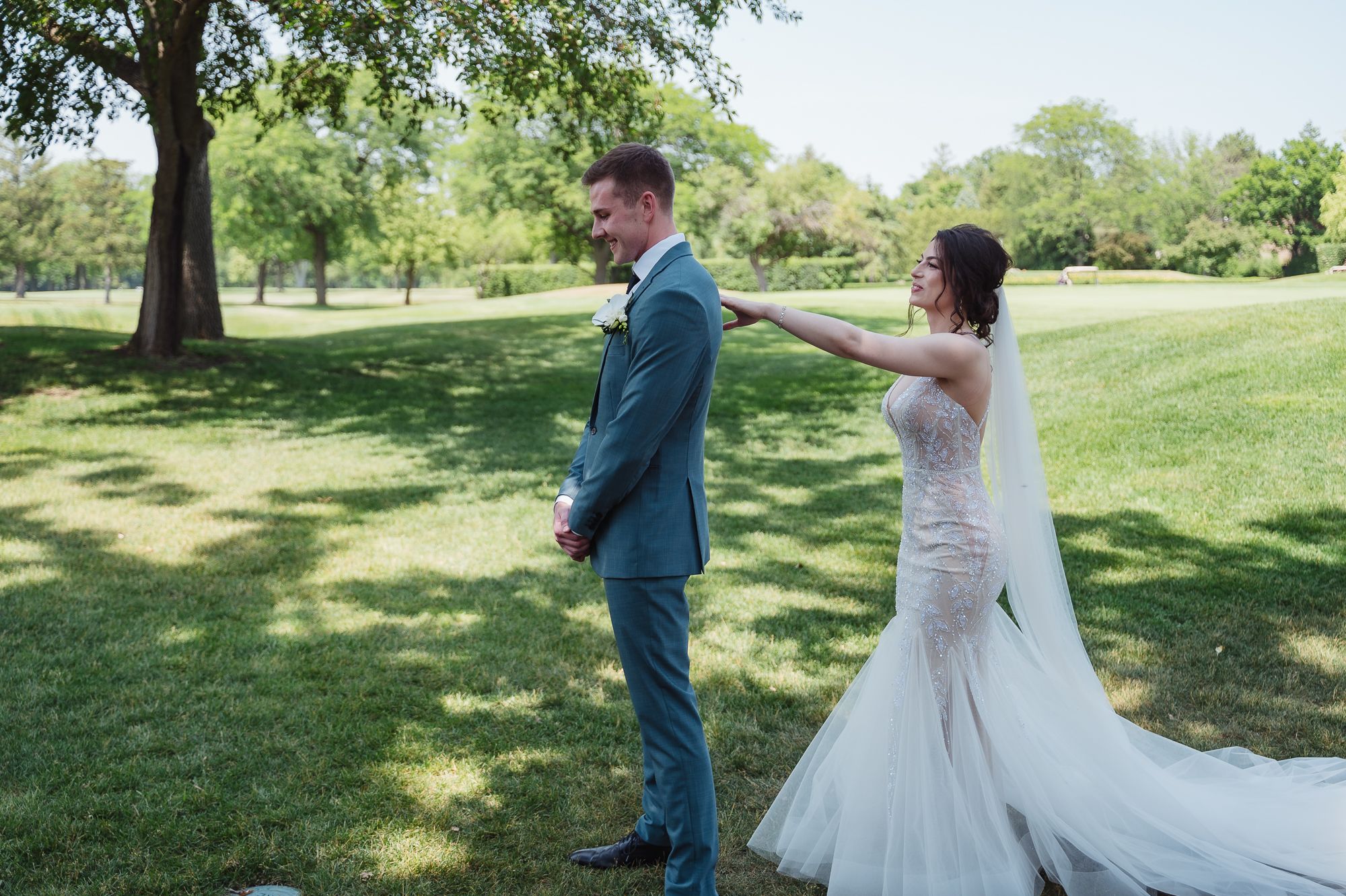 Bride in lace mermaid gown taps groom's shoulder during first look on green lawn.