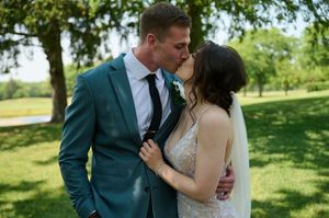 Bride and groom sharing a kiss outdoors in a lush green park, groom in teal suit, bride in lace wedding gown.
