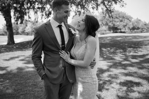 Black and white photo of a smiling couple in wedding attire embracing outdoors under trees.
