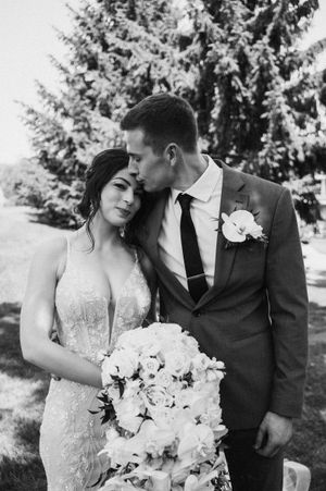 Groom kissing bride on temple while she holds white floral bouquet, elegant wedding portrait in black and white.