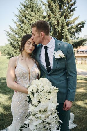 Bride in beaded gown holds white cascading bouquet as groom in teal suit kisses her forehead outdoors.