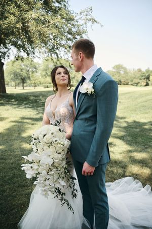 Bride holding white cascading bouquet gazes at groom in blue suit outdoors on lush green lawn.