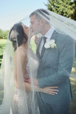 Bride and groom sharing a romantic kiss under a flowing white veil outdoors on their wedding day.