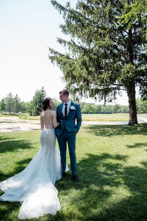 Bride in lace gown and groom in teal suit share a romantic moment on green lawn beside tall evergreen tree.