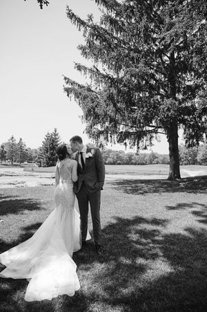 Black and white photo of a bride and groom sharing a kiss outdoors near a large evergreen tree on their wedding day.