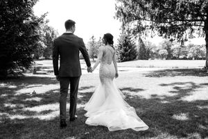 Black and white photo of a bride and groom holding hands, walking through a sunny park, wedding dress with elegant train.