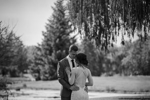 Black and white photo of a bride and groom embracing outdoors near a weeping willow tree and pond.