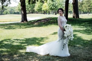 Bride in a lace mermaid wedding gown holding a white floral bouquet, posing outdoors in a lush green park.