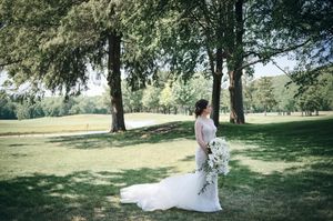 Bride in white gown with long train holds bouquet while standing under trees in lush green park setting.