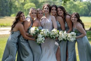 Bride in lace gown poses with six bridesmaids in sage green dresses holding white floral bouquets outdoors.