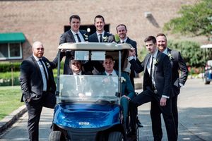 Groomsmen in black suits posing with a blue golf cart outdoors at a wedding venue on a sunny day.