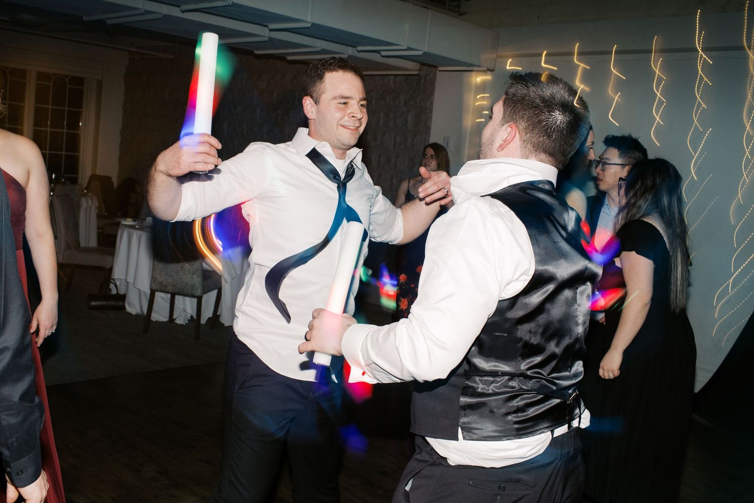 Two men dancing with glowing LED light sticks at a lively wedding reception party.