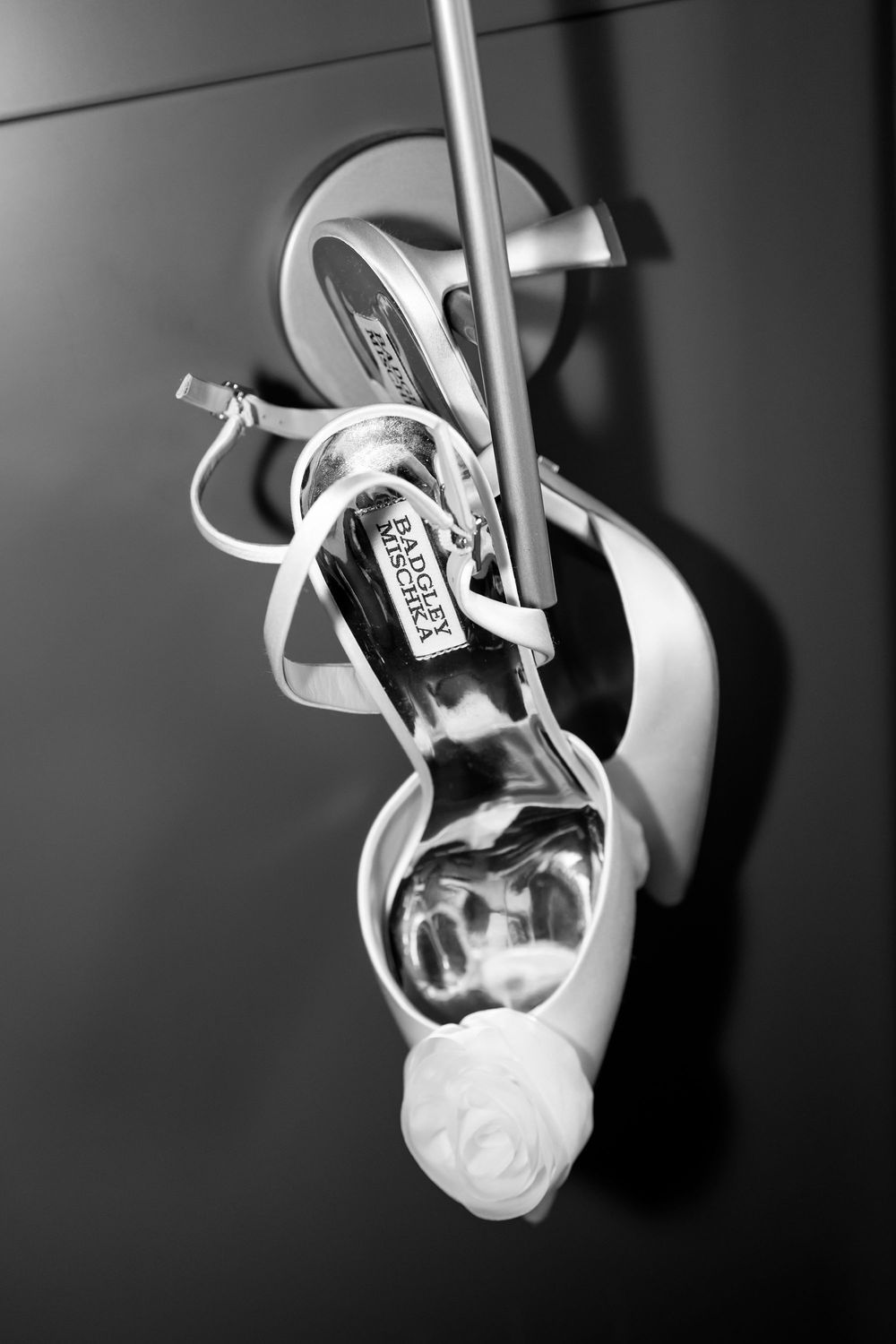 Black and white photo of Badgley Mischka bridal heels with rose detail hanging elegantly.