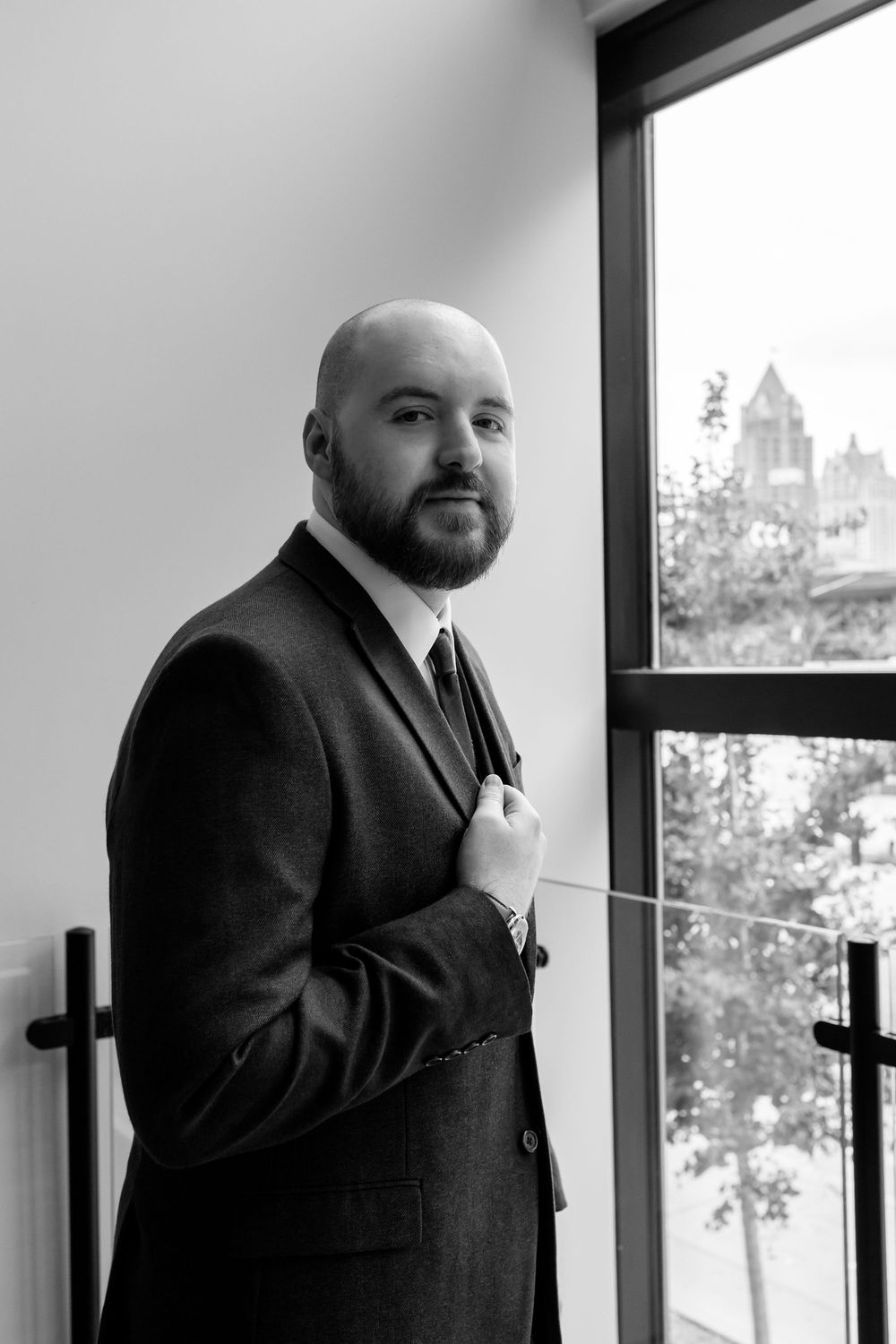 Black and white portrait of a bald bearded man in a suit adjusting his jacket near a large window.