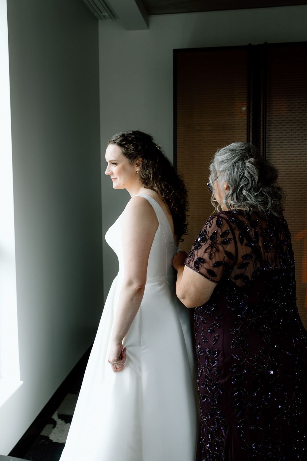 Bride in white gown stands by window as older woman in beaded burgundy dress helps with final wedding preparations.