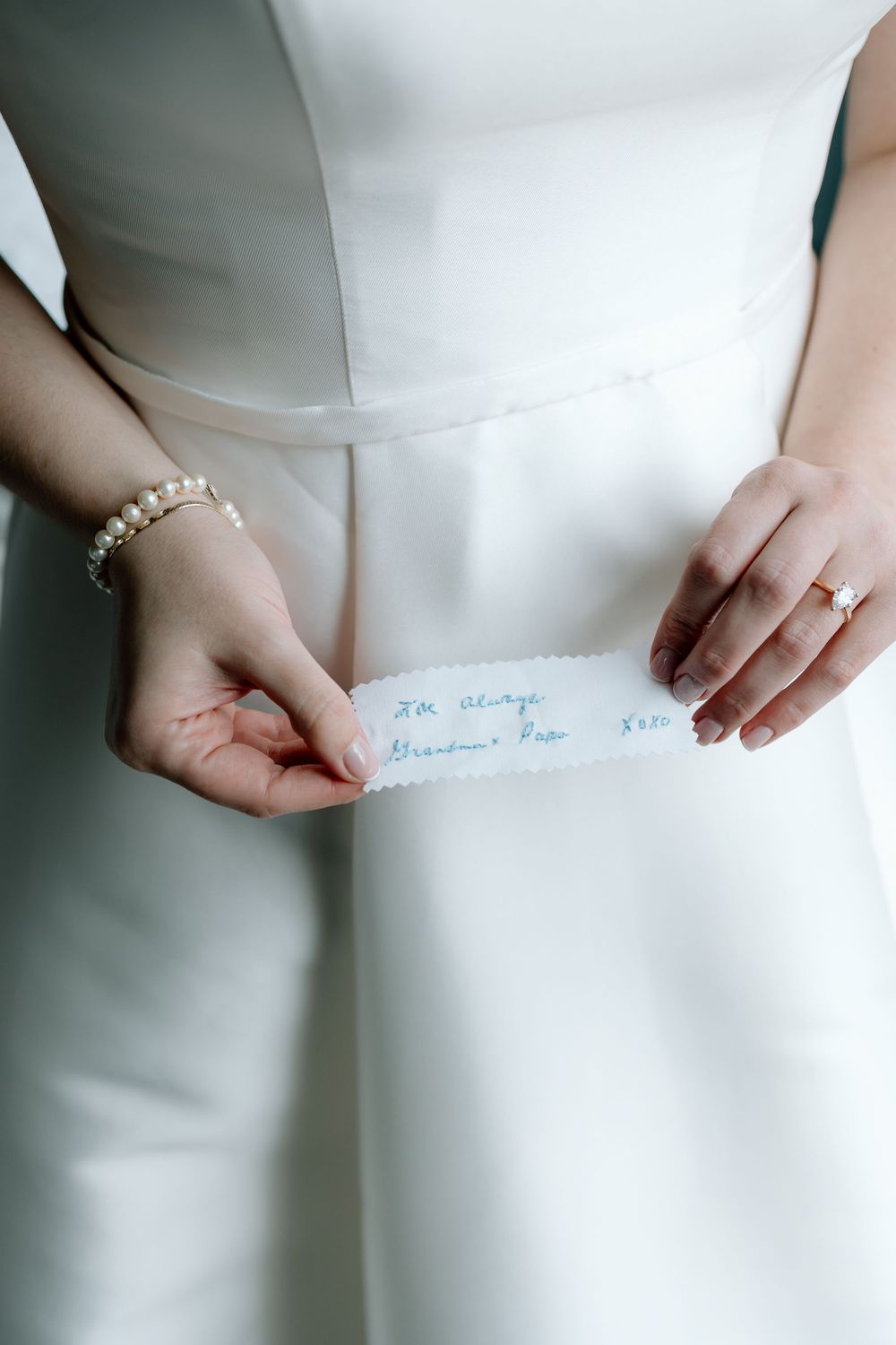 Bride in white gown holds a handwritten note with pearl bracelet and engagement ring.