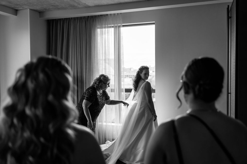 Black and white photo of a bride getting her wedding dress adjusted by a helper near a bright window.