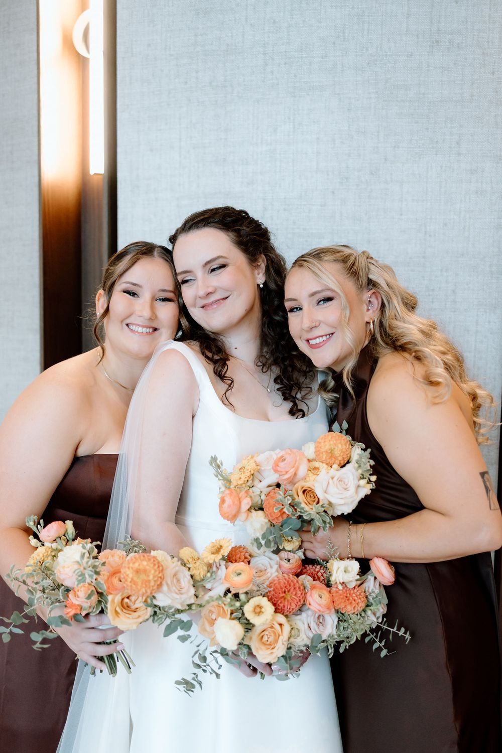 Bride poses with two bridesmaids in brown dresses holding peach and orange floral bouquets before the wedding ceremony.