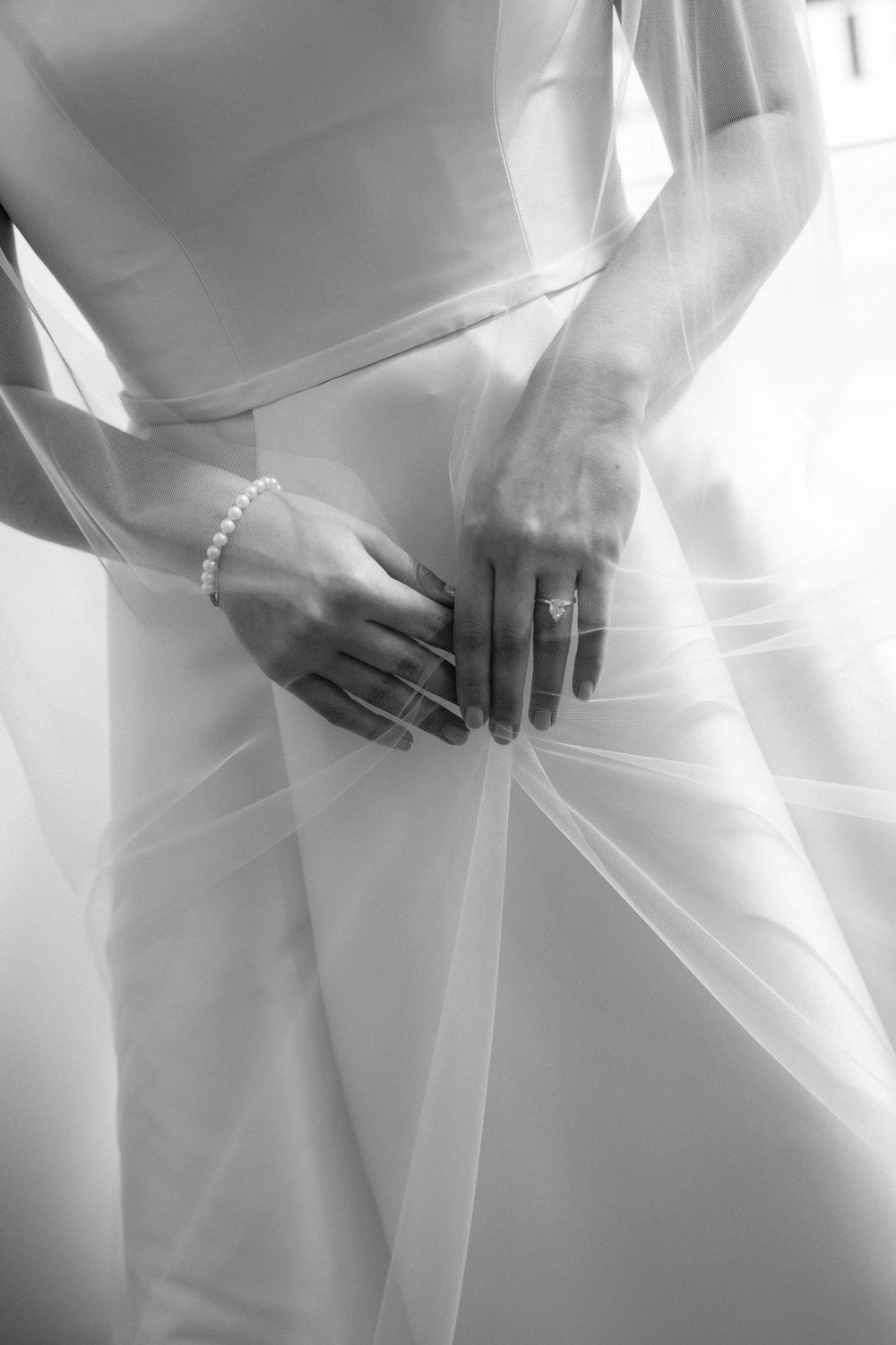 Black and white close-up of a bride holding her wedding dress, wearing a diamond ring and pearl bracelet.