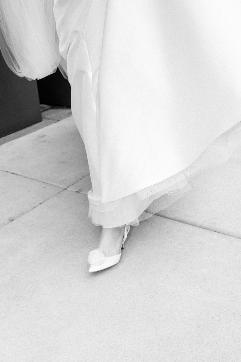 Black and white photo of a bride walking in a flowing white wedding gown and elegant heels on stone pavement.