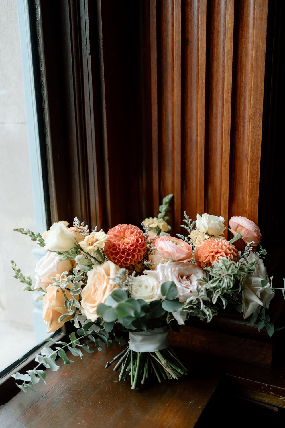 Lush bridal bouquet featuring coral dahlias, peach roses, and eucalyptus displayed on a wooden surface by a window.