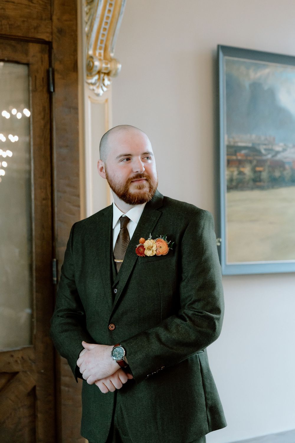 Groom in green tweed suit with floral boutonniere standing near ornate door at wedding venue.