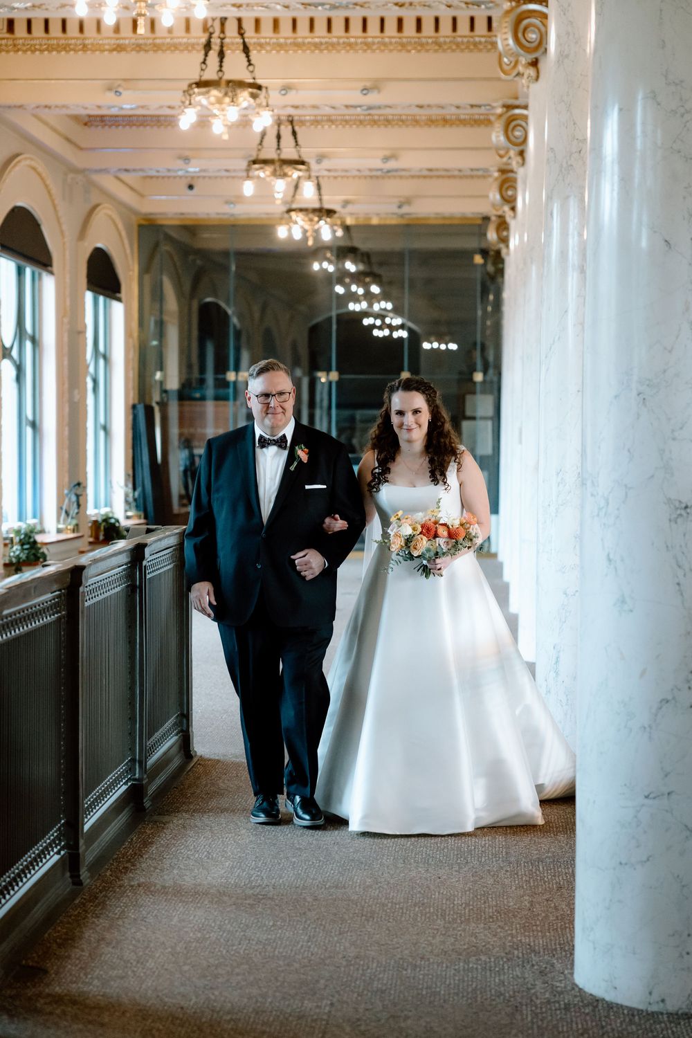 Bride in white ball gown walks down elegant venue hallway with older man in tuxedo, holding colorful bouquet.