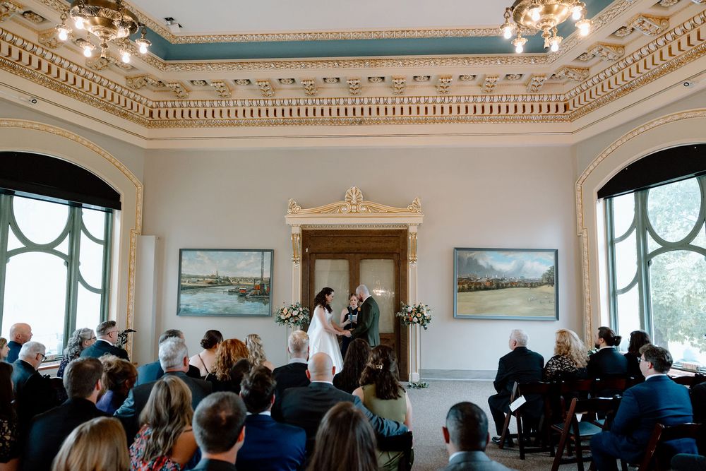 Elegant wedding ceremony in an ornate room with guests seated as couple exchanges vows at the altar.